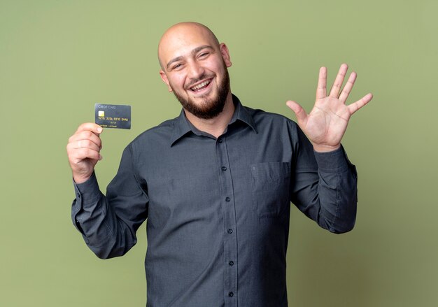 クレジットカードを保持し、オリーブグリーンで隔離の手で5を示すうれしそうな若いハゲのコールセンターの男