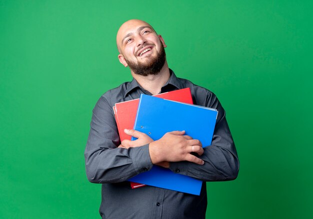 Joyful young bald call center man holding folders looking up isolated on green