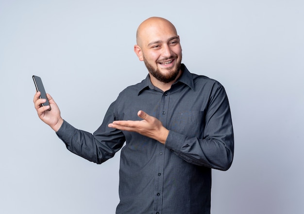 Joyful young bald call center man holding mobile phone looking at side and showing empty hand isolated on white