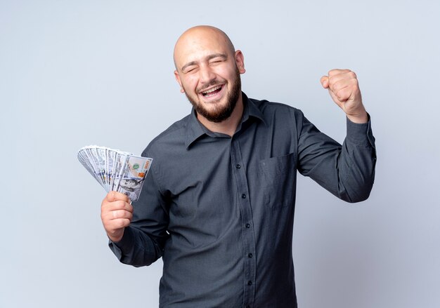 Joyful young bald call center man holding money and clenching fist with closed eyes isolated on white