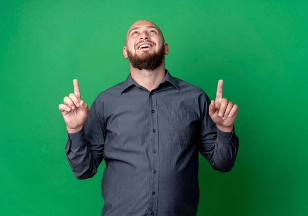 Joyful young bald call center man looking and pointing up isolated on green