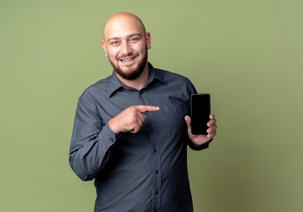 Радостный молодой лысый человек из колл-центра показывает держа и указывая на мобильный телефон, изолированный на оливково-зеленом