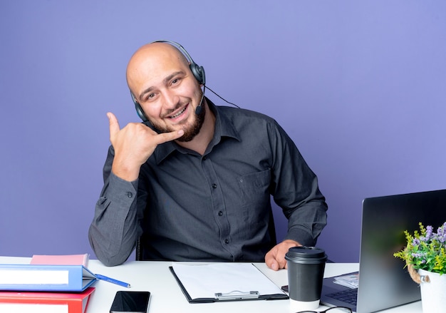 Joyful young bald call center man wearing headset sitting at desk with work tools doing call gesture isolated on purple