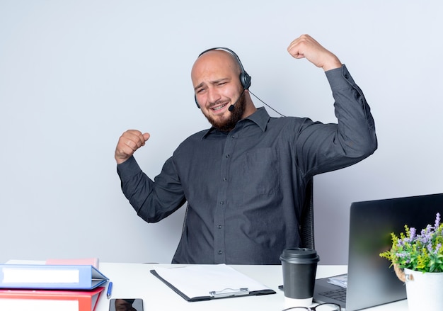 Joyful young bald call center man wearing headset sitting at desk with work tools gesturing strong isolated on white