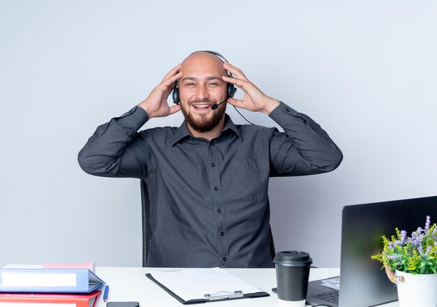 Joyful young bald call center man wearing headset sitting at desk with work tools putting hands on headset isolated on white