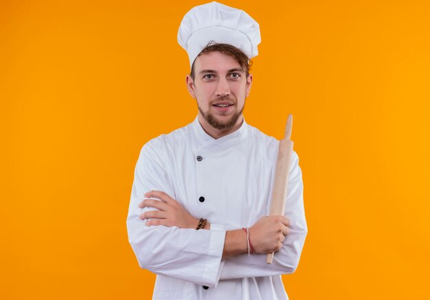 A joyful young bearded chef man in white uniform holding rolling pin with folded hands while looking on an orange wall