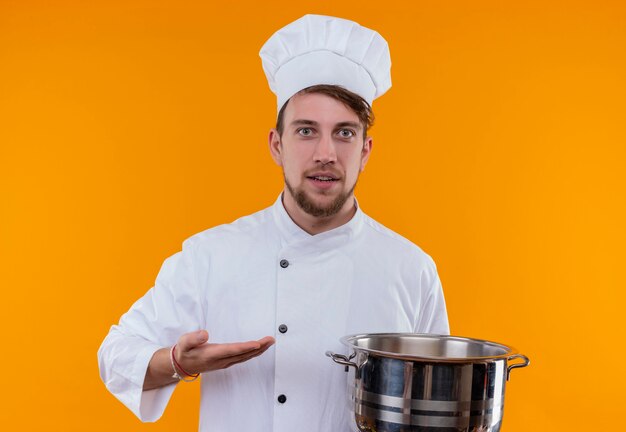 A joyful young bearded chef man in white uniform showing sauce pan while looking on an orange wall