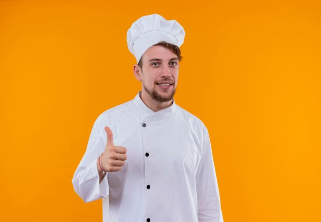 A joyful young bearded chef man in white uniform showing thumbs up while looking on an orange wall