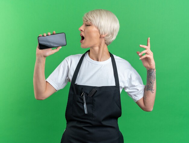 Joyful young beautiful female barber in uniform holding phone and singing isolated on green wall