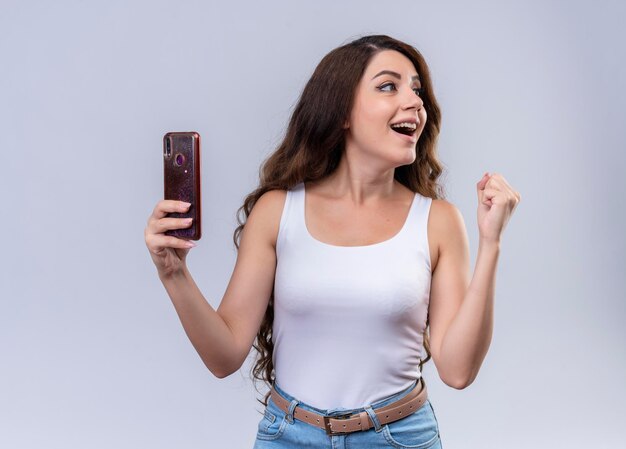 Joyful young beautiful girl holding mobile phone looking at it and raising fist 