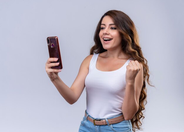 Joyful young beautiful girl holding mobile phone looking at it and raising fist 