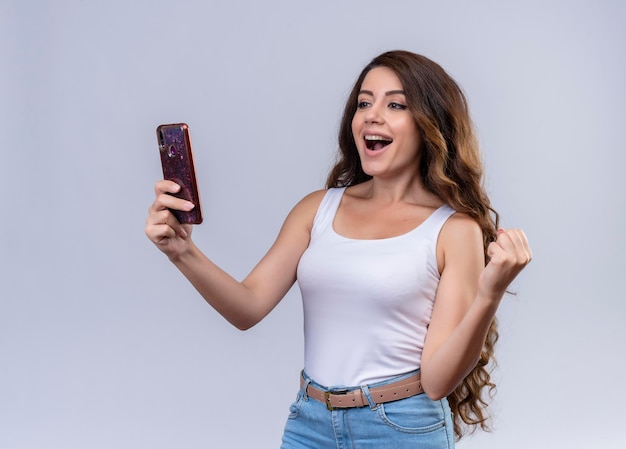 Joyful young beautiful girl holding mobile phone and looking at it with raised fist 