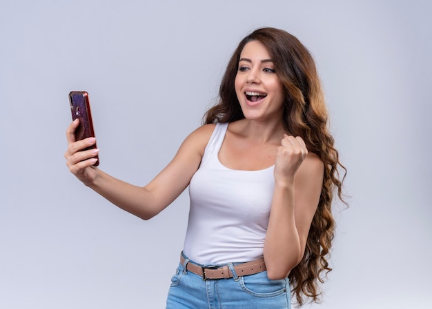 Joyful young beautiful girl holding mobile phone looking at it with raised hand 