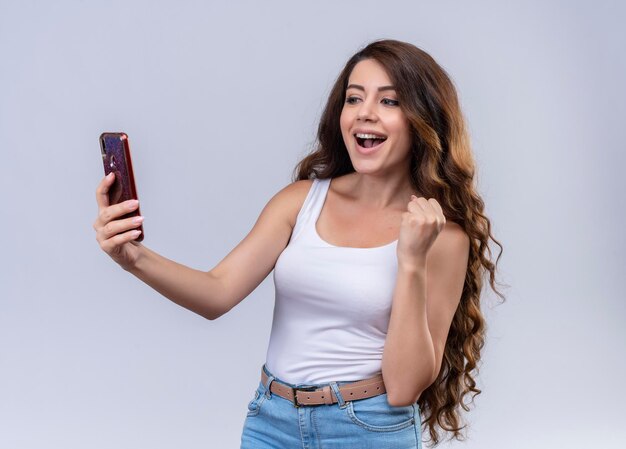 Joyful young beautiful girl holding mobile phone looking at it with raised hand 