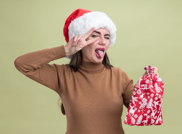 올리브 녹색 벽에 고립 된 혀와 평화 제스처를 보여주는 크리스마스 가방을 들고 크리스마스 모자를 쓰고 즐거운 젊은 아름 다운 소녀