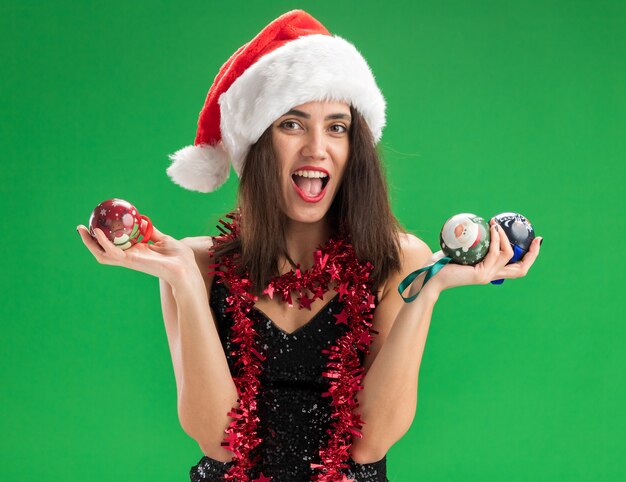 Joyful young beautiful girl wearing christmas hat with garland on neck holding christmas tree balls isolated on green wall