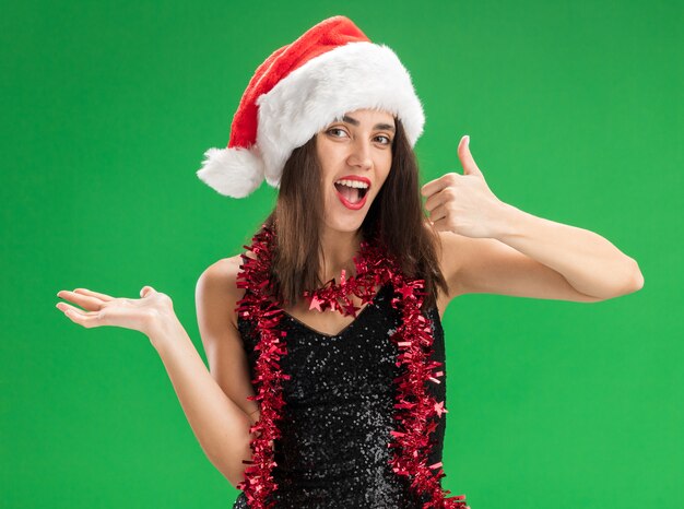 Joyful young beautiful girl wearing christmas hat with garland on neck showing thumb up spreading hand isolated on green background