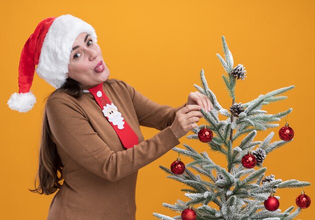Радостная молодая красивая девушка в новогодней шапке с галстуком стоит рядом с елкой, показывая язык на оранжевом фоне