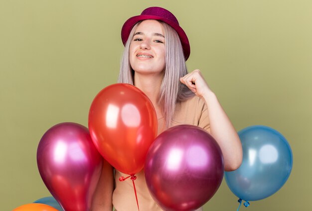 Joyful young beautiful girl wearing dental braces with party hat standing behind balloons showing yes gesture 