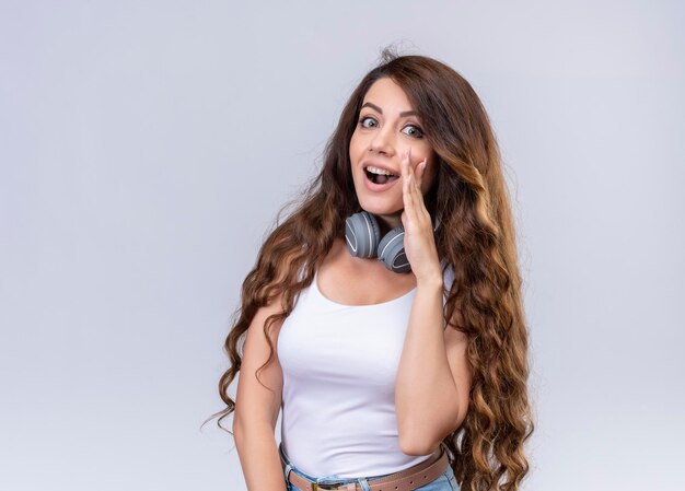 Joyful young beautiful girl wearing headphones on neck putting hand near mouth  with copy space