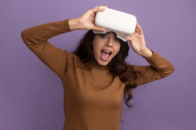 紫の壁に分離された vr ヘッドセットを着て保持しているうれしそうな若い美しい女の子