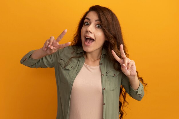 Joyful young beautiful girl wearing olive green t-shirt showing peace gesture isolated on yellow wall