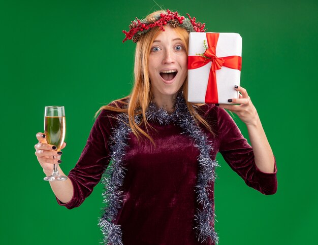 Joyful young beautiful girl wearing red dress with wreath and garland on neck holding glass of champagne with gift box isolated on green wall