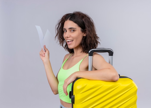 Joyful young beautiful traveler woman holding airplane tickets and suitcase on isolated white wall