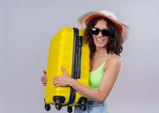 Joyful young beautiful traveler woman wearing hat and sunglasses and holding suitcase on isolated white wall