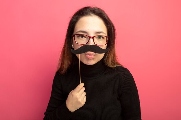 Joyful young beautiful woman in a black turtleneck and glasses holding funny mustache on stick standing over pink wall