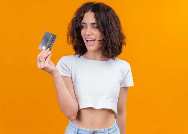 Joyful young beautiful woman holding card and looking at it on isolated orange wall with copy space