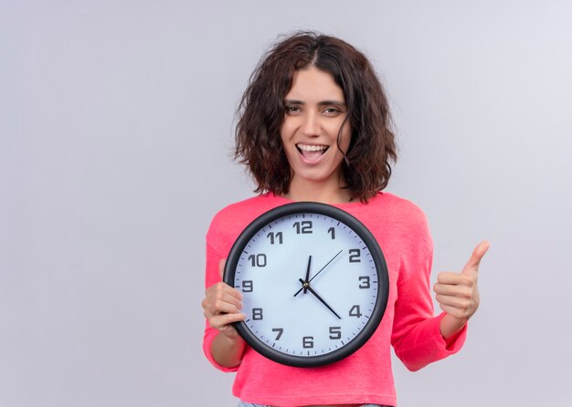 Joyful young beautiful woman holding clock and showing thumb up on isolated white wall with copy space