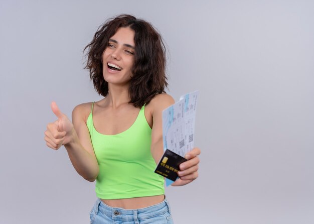 Joyful young beautiful woman stretching airplane tickets and card and showing thumb up on isolated white wall with copy space