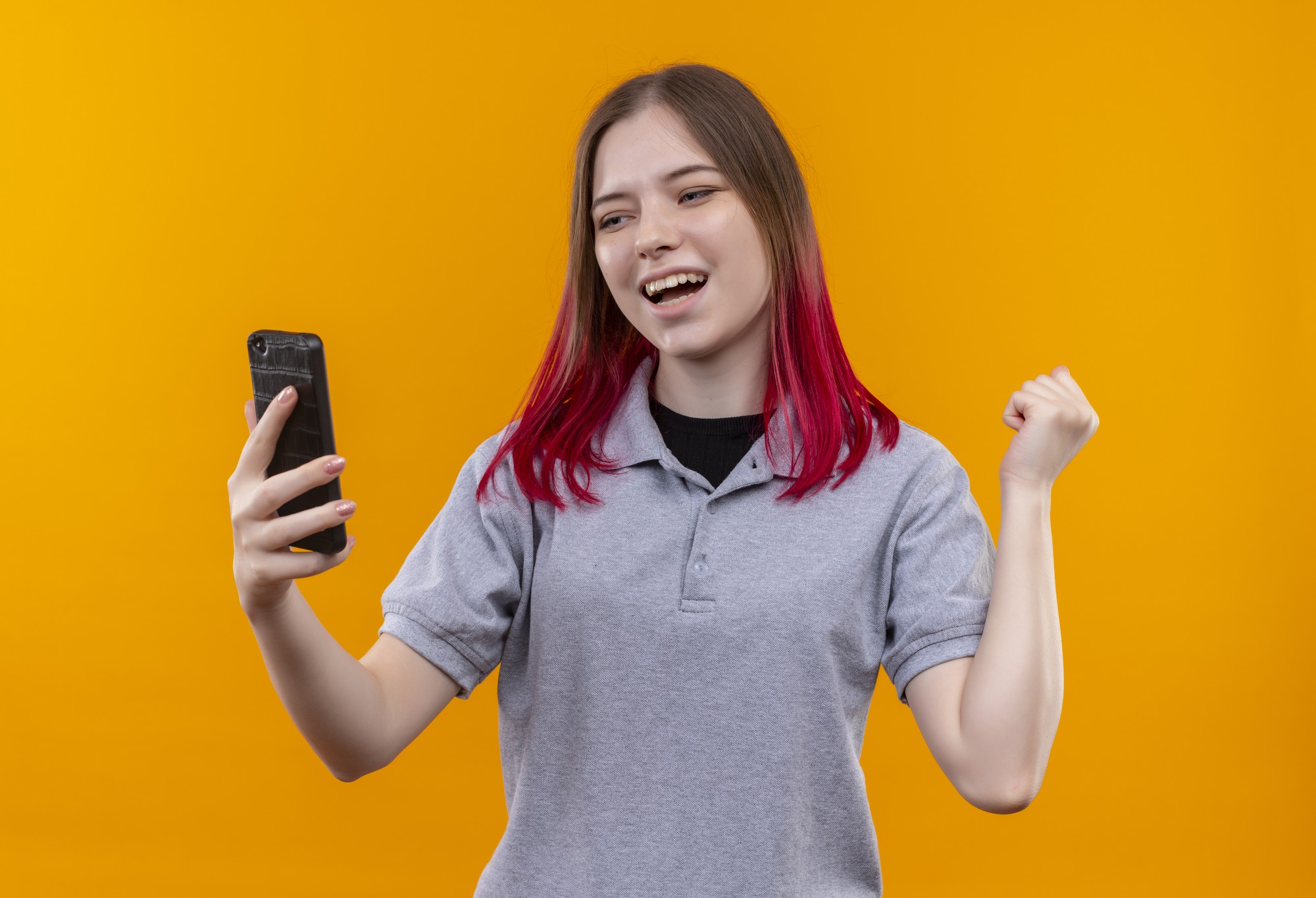 Joyful young beautiful woman wearing gray t-shirt looking at phone in her hand showing yes gesture on isolated yellow wall