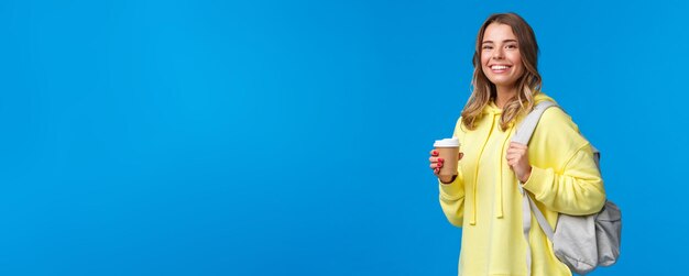 Joyful young blond female student with backpack smiling camera satisfied as holding cup of takeaway