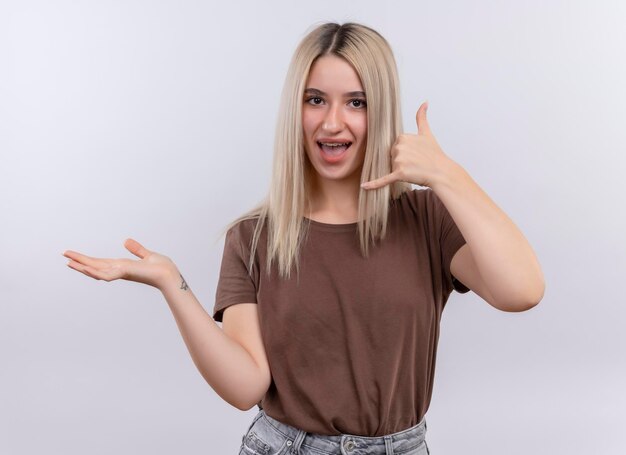 Joyful young blonde girl in dental braces doing call gesture and showing empty hand on isolated white space