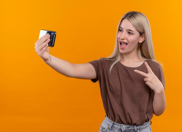 Joyful young blonde girl in dental braces doing peace sign and taking selfie on isolated orange space