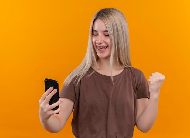 Радостная молодая блондинка в брекетах держит мобильный телефон и смотрит на него с поднятым кулаком на изолированном оранжевом пространстве