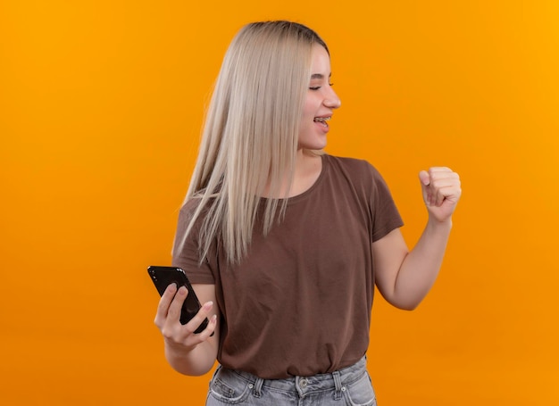 Joyful young blonde girl in dental braces holding mobile phone with raised fist on isolated orange space with copy space
