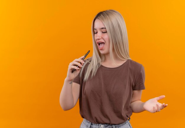 Joyful young blonde girl in dental braces singing using phone as microphone showing empty hand with closed eyes on isolated orange space with copy space