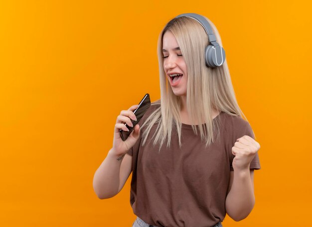 Радостная молодая блондинка в брекетах в наушниках поет, используя телефон в качестве микрофона с поднятой рукой и закрытыми глазами на изолированном оранжевом пространстве с копией пространства