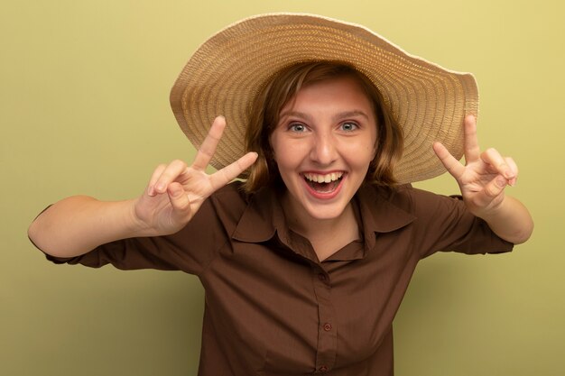 オリーブグリーンの壁に分離された平和のサインをやってビーチ帽子をかぶってうれしそうな若いブロンドの女の子