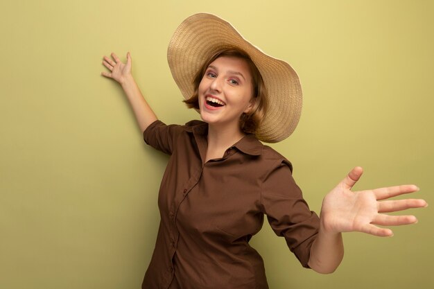 Joyful young blonde girl wearing beach hat stretching out hands looking  isolated on olive green wall with copy space