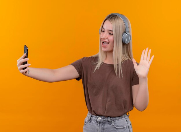 Радостная молодая блондинка в наушниках в брекетах держит мобильный телефон и машет ему на изолированном оранжевом пространстве
