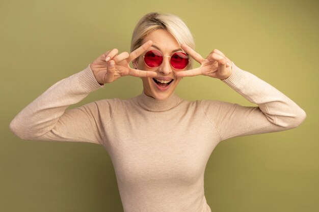 Joyful young blonde girl wearing sunglasses showing v-sign symbols near eyes 