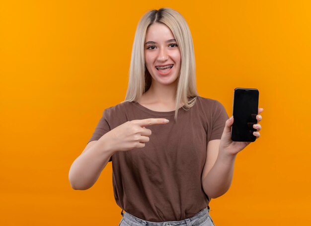Радостная молодая блондинка с брекетами держит мобильный телефон и указывает на него на изолированном оранжевом пространстве с копией пространства