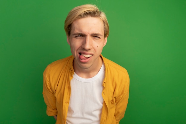 Joyful young blonde guy wearing yellow t-shirt showing tongue isolated on green with copy space