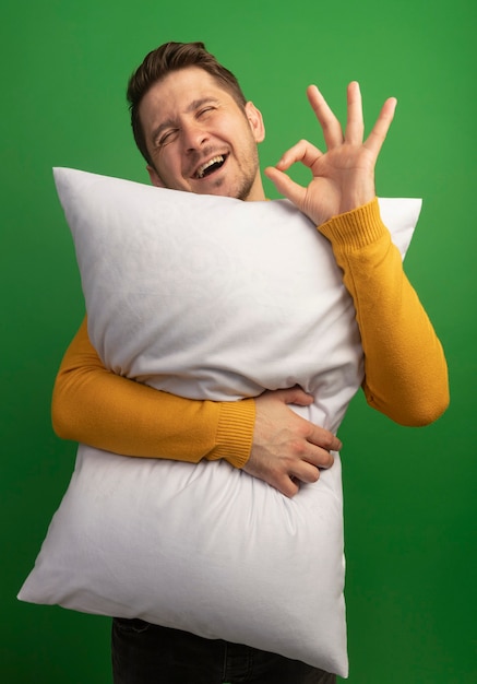 OKサインをしている枕を抱き締めるうれしそうな若いブロンドのハンサムな男