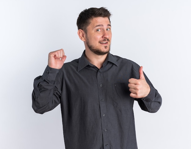 Joyful young blonde handsome man showing fist and thumb up looking at camera 