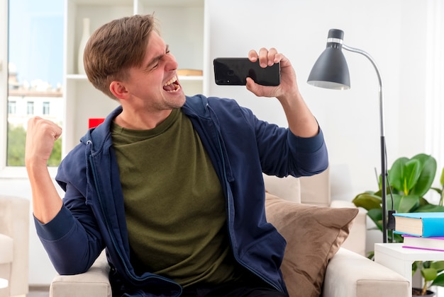 Joyful young blonde handsome man sits on armchair keeping fist and holding phone